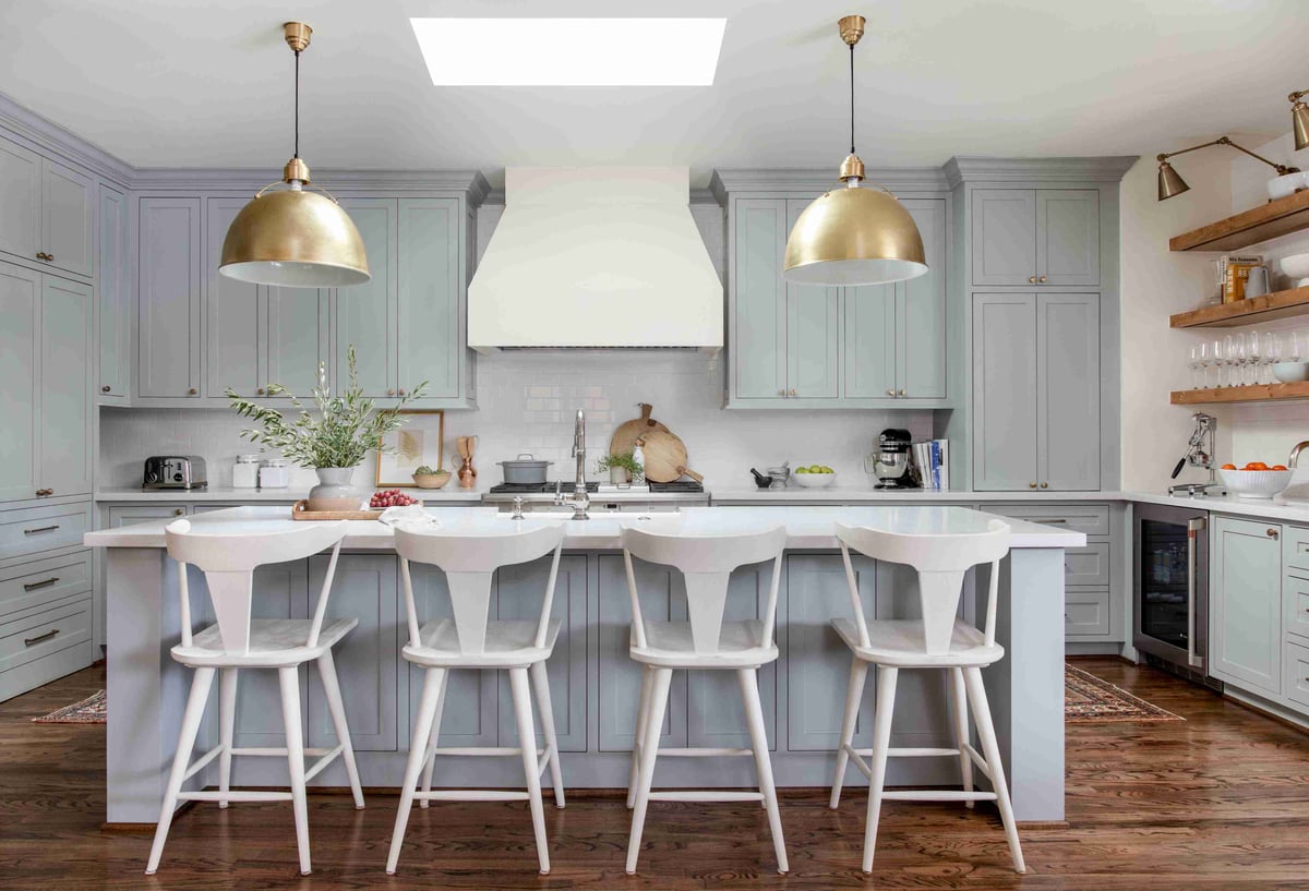 A light-filled kitchen with pastel blue cabinets, white countertops, two large gold pendant lights.